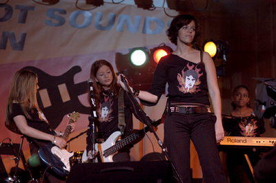 On-Screen Performance – Hell Hath No Fury (2003)
Still image from Season 3, Episode 18 (“Rock and Roll High”) of Degrassi: The Next Generation, showing Ashley, Ellie, Paige, and Hazel performing as the all-girl band Hell Hath No Fury at the school’s Battle of the Bands. The girls wore matching tees featuring the band logo, which were later sold in the 2007 official Degrassi Online Store. Their high-energy performance was aimed at Craig’s band, Downtown Sasquatch — proving that these girls meant business.
Keywords: Degrassi;Hell Hath No Fury;Rock and Roll High;Ashley Kerwin;Paige Michalchuk;Ellie Nash;Hazel Aden;Band Performance;Downtown Sasquatch;Season 3;2003